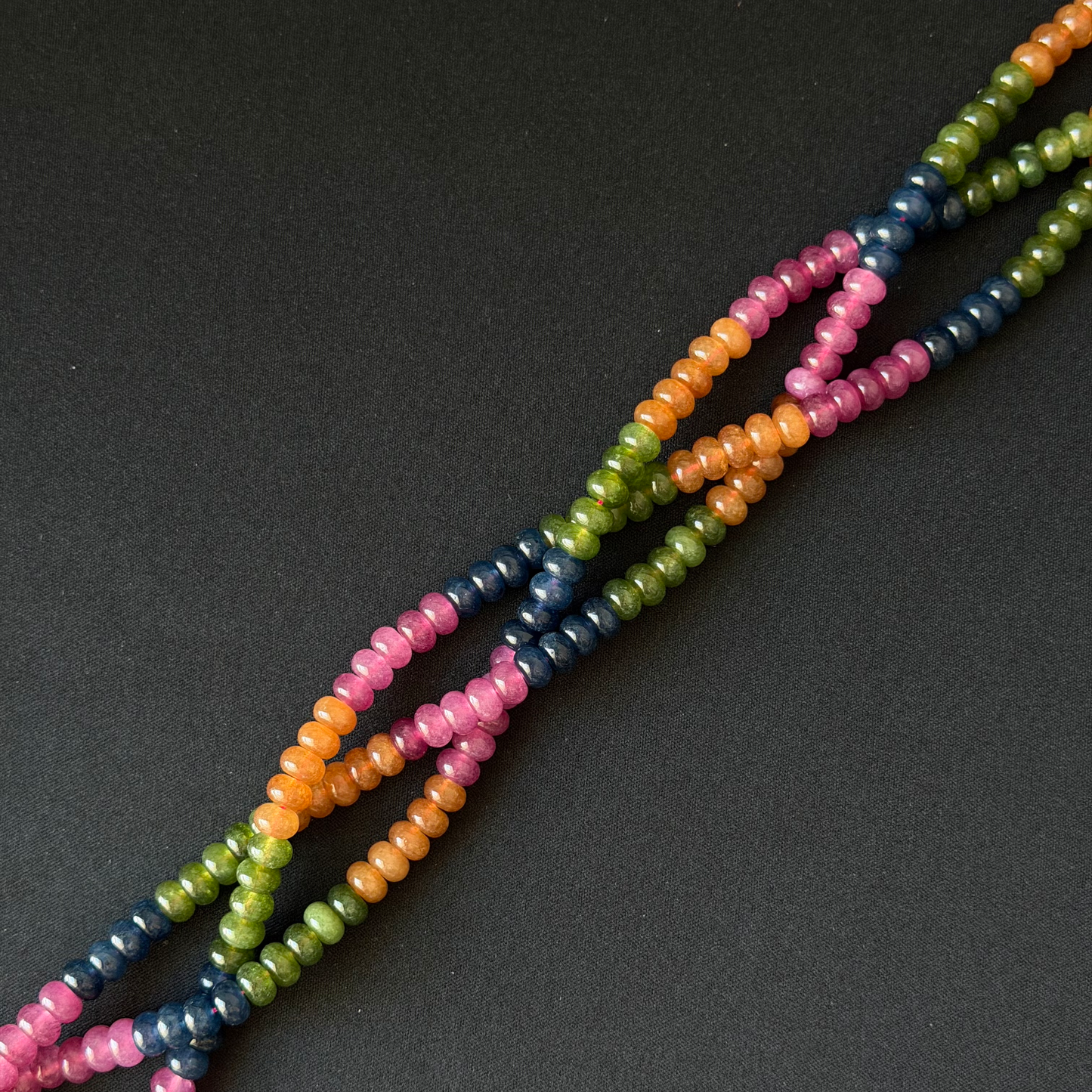 Multicolor tourmaline dyed jade beaded strands with small rondelle beads in pink, orange, green, and navy blue, arranged diagonally on a dark gray background.