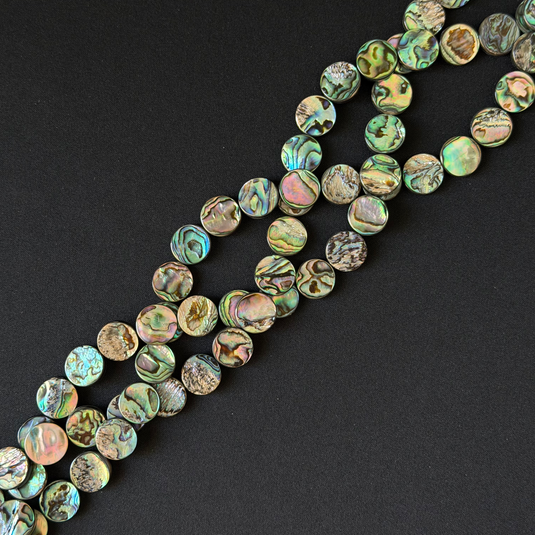 Round coin shape abalone shell beads with iridescent green, blue, and brown patterns, arranged diagonally on a black background.