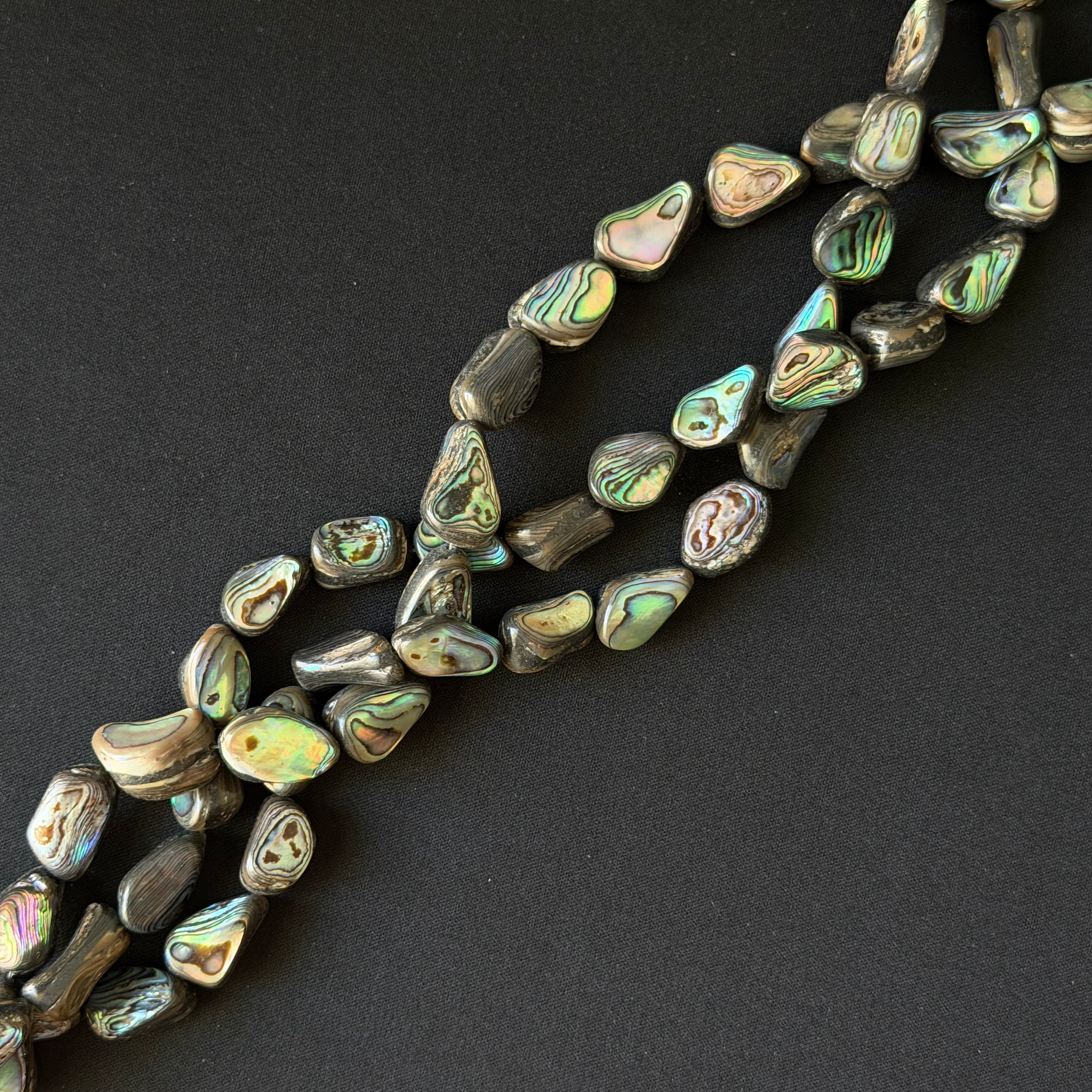 Tumbled abalone shell beads with natural iridescent colors, arranged in diagonal strands on a dark textured background.