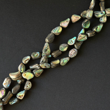 Tumbled abalone shell beads with natural iridescent colors, arranged in diagonal strands on a dark textured background.