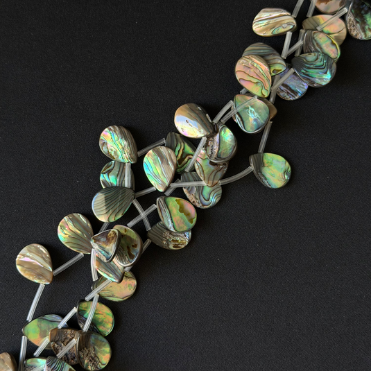 Abalone shell bead strand on a black background, featuring iridescent green, blue, and brown shell pieces connected by gray string. teardrop shape