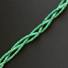 A close-up of a strand of small cylindrical heishi beads in a turquoise green color with natural matrix inclusions, arranged diagonally on a dark gray background.