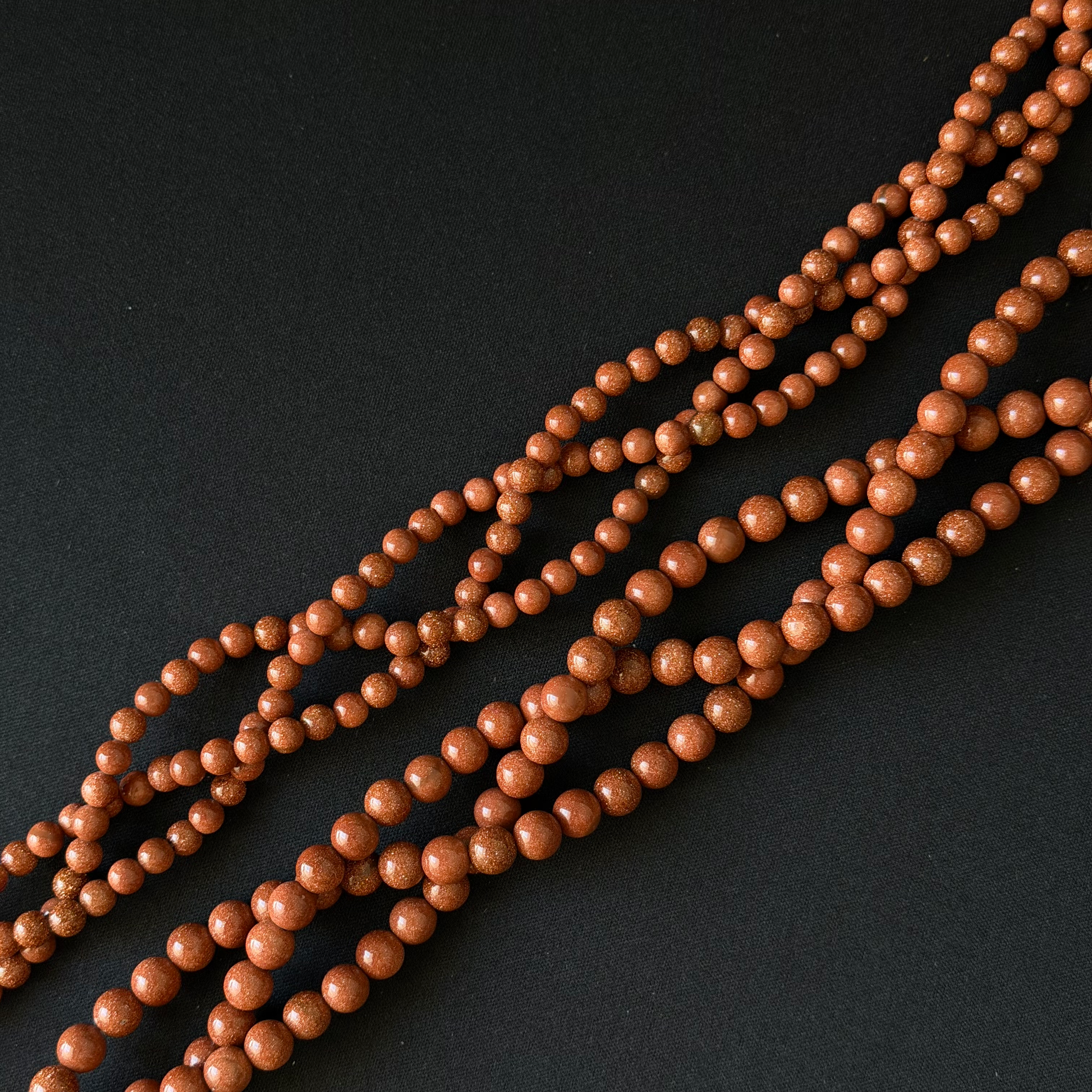Multiple strands of small, polished round beads in a warm reddish-brown or terracotta color, arranged diagonally on a dark black background.