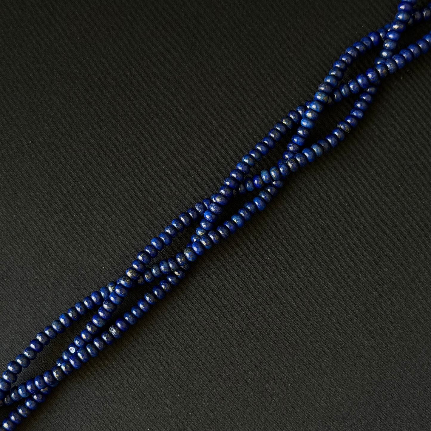 A close-up of a strand of small, dark blue smooth dyed lapis rondelle beads arranged diagonally on a dark, textured background.
