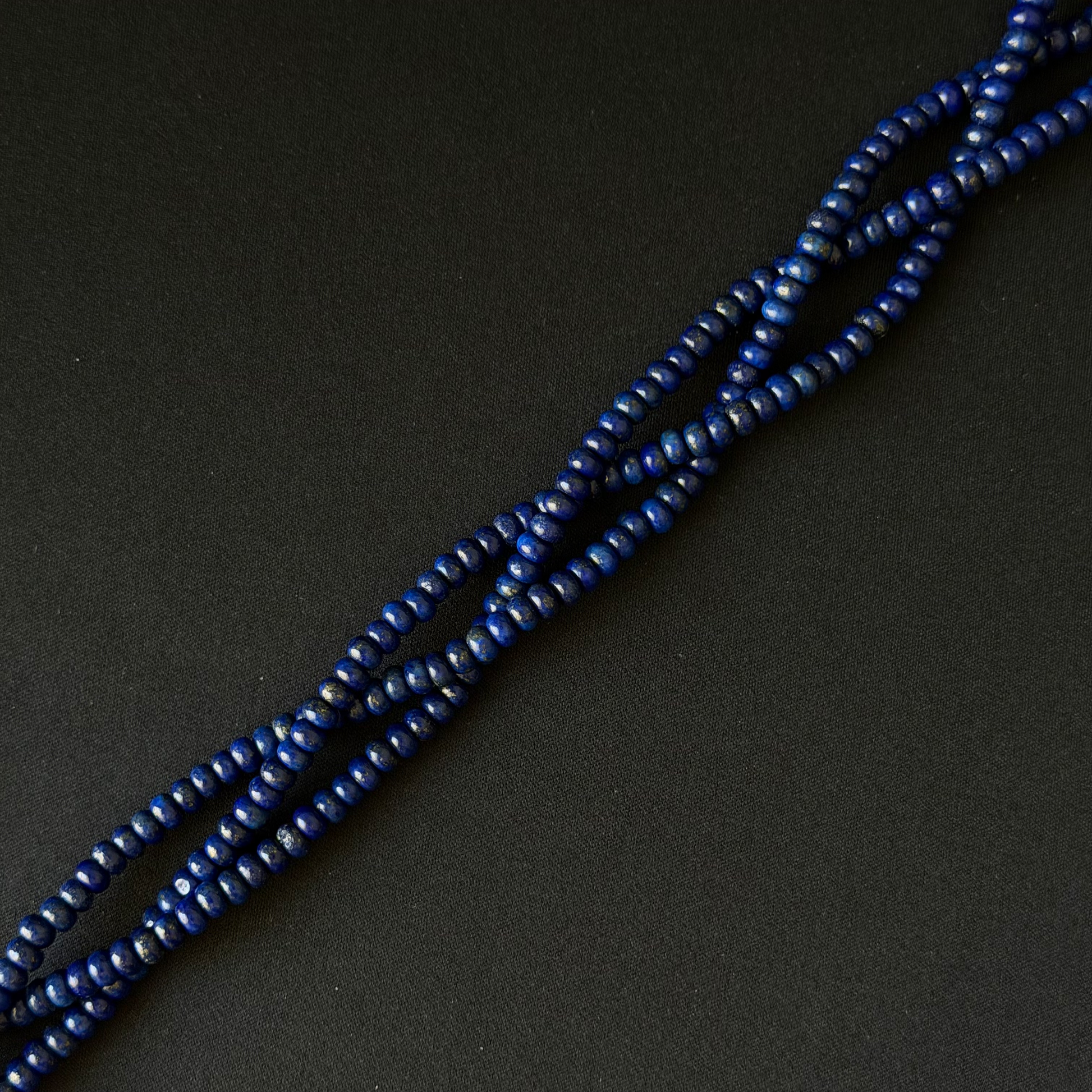 A close-up of a strand of small, dark blue smooth dyed lapis rondelle beads arranged diagonally on a dark, textured background.