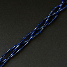 A close-up of a strand of small, dark blue smooth dyed lapis rondelle beads arranged diagonally on a dark, textured background.