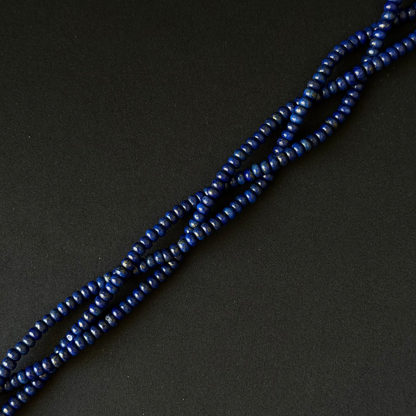 A close-up of a strand of small, dark blue smooth dyed lapis rondelle beads arranged diagonally on a dark, textured background.