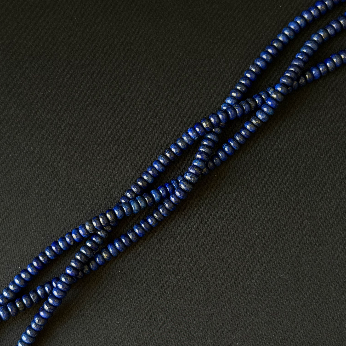 Three strands of small dark blue dyed lapis lazuli beads arranged diagonally on a dark gray background.