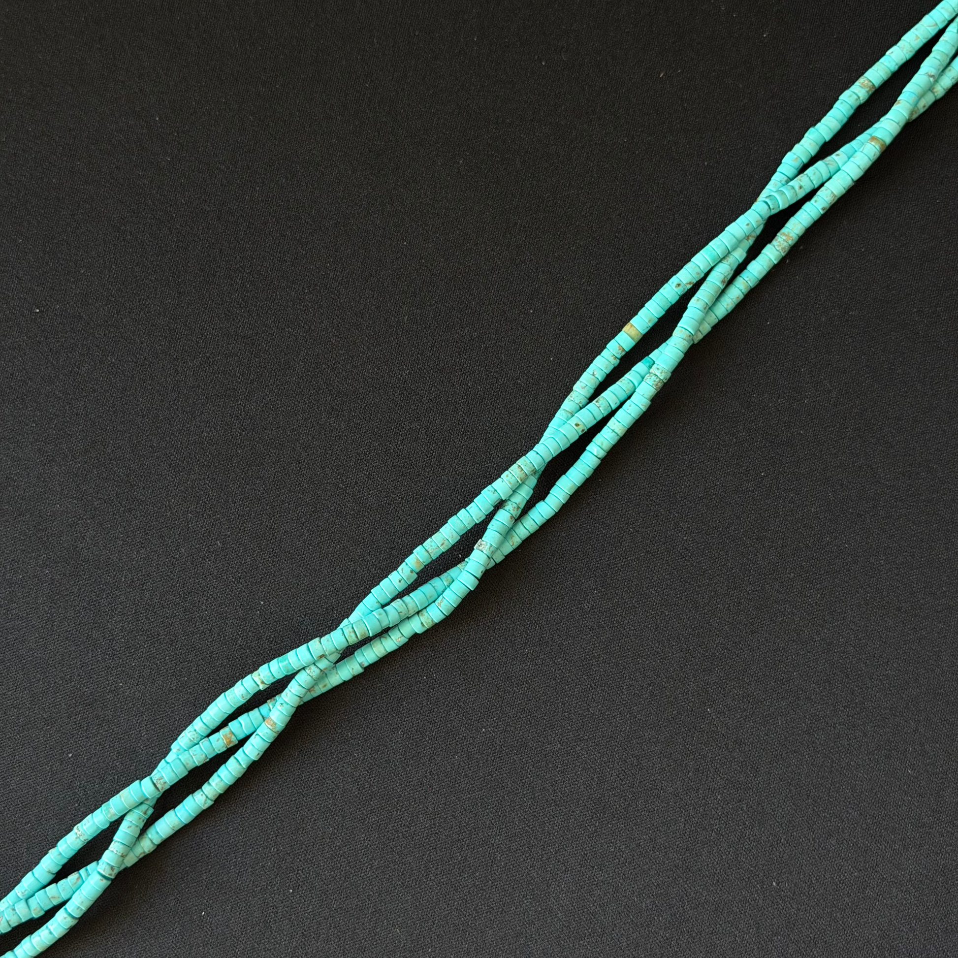 A strand of small turquoise-blue tube shape beads strung diagonally on a dark black background.