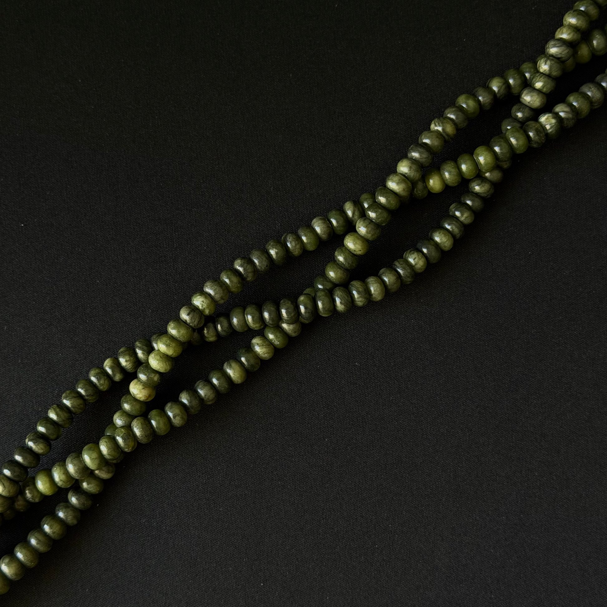 A close-up of olive green smooth rondelle beads strung on a cord, arranged diagonally on a dark black background.