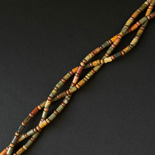 A close-up of a multi-strand beads for necklace or bracelet made of cylindrical beads in earthy olive green, terracotta red, ochre yellow, and brown tones, arranged diagonally on a dark textured background.