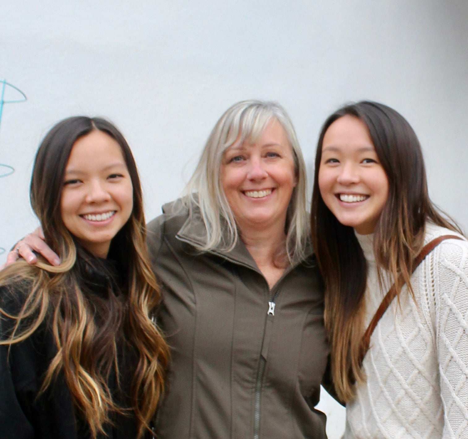 Three women smiling and posing together outdoors against a light gray wall. The woman in the center wears an olive green zip-up jacket, while the woman on the right wears a white cable-knit sweater.