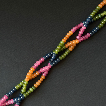 Multicolor tourmaline dyed jade beaded strands with small rondelle beads in pink, orange, green, and navy blue, arranged diagonally on a dark gray background.