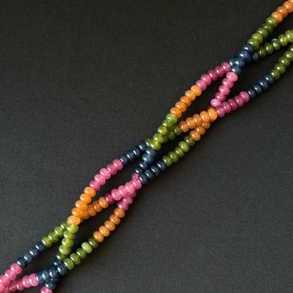 Multicolor tourmaline dyed jade beaded strands with small rondelle beads in pink, orange, green, and navy blue, arranged diagonally on a dark gray background.