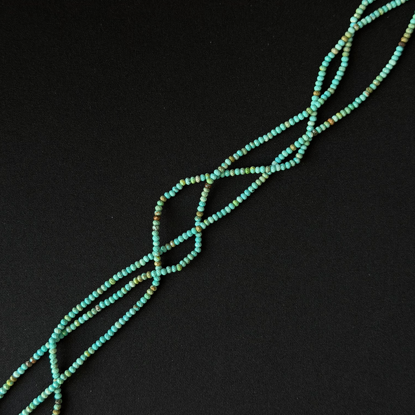 A close-up of a beaded strand or necklace made of small turquoise-blue beads with subtle multicolor flecks, arranged diagonally on a solid black background.