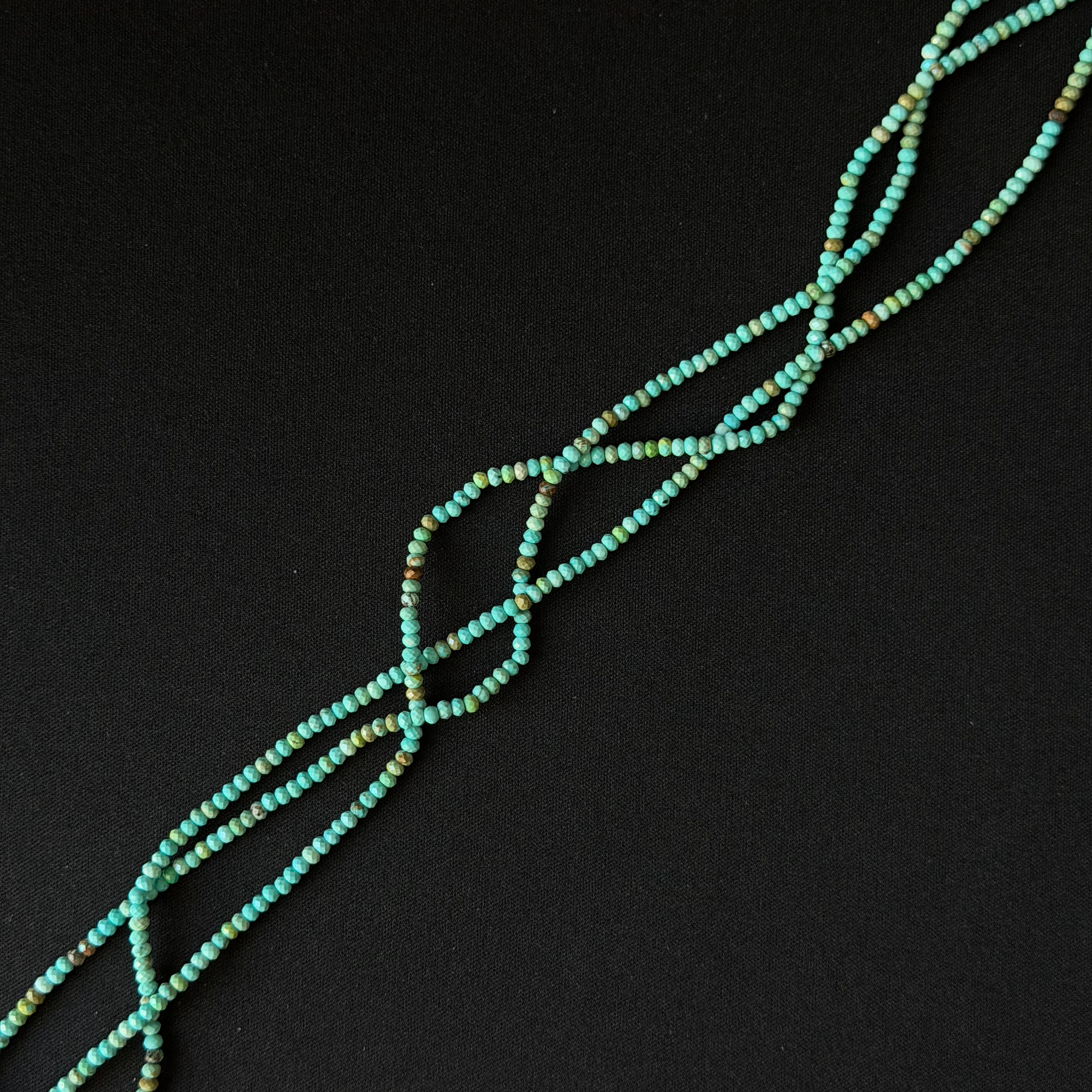 A close-up of a beaded strand or necklace made of small turquoise-blue beads with subtle multicolor flecks, arranged diagonally on a solid black background.