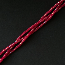 A close-up of several strands of faceted, seed-like beads in a deep red or burgundy tone, arranged diagonally on a dark black background.