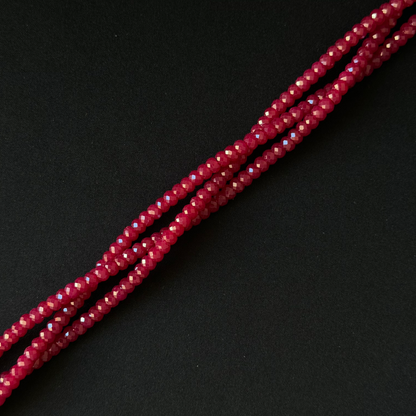 A close-up of several strands of faceted, seed-like beads in a deep red or burgundy tone, arranged diagonally on a dark black background.