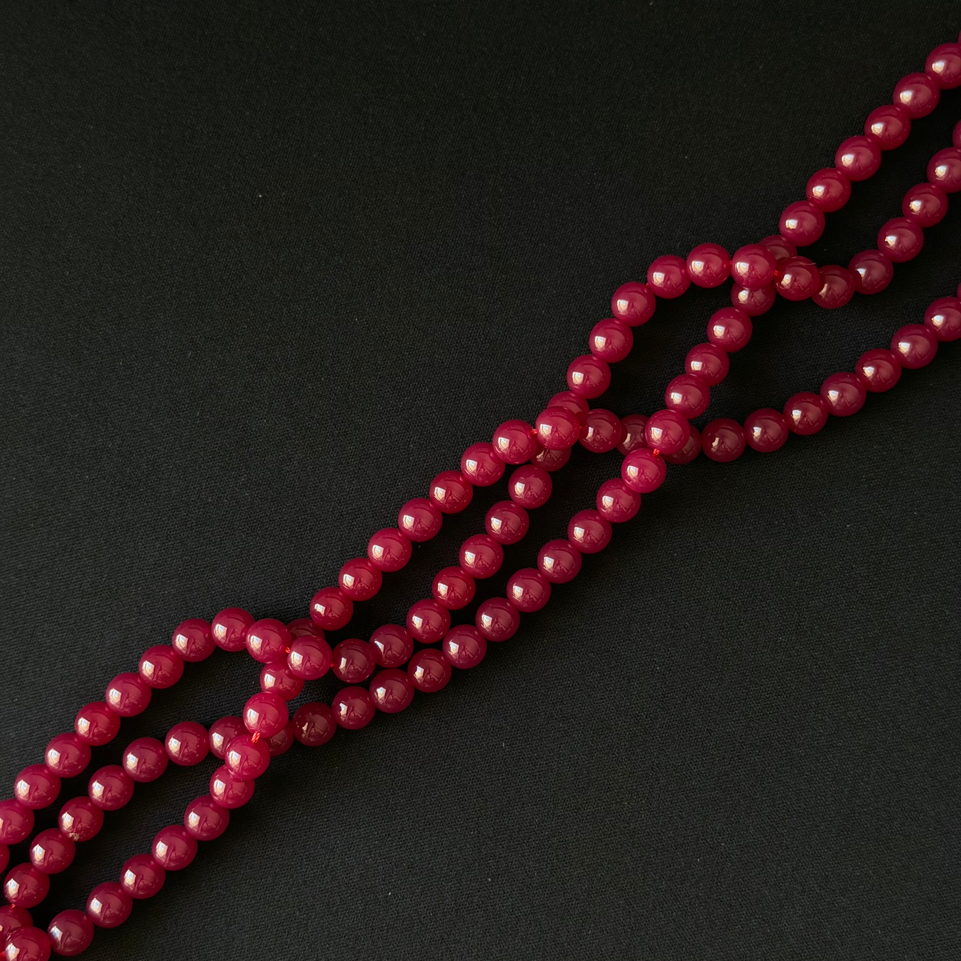 A close-up of a strand of faceted, round beads in a deep magenta-pink color, arranged diagonally on a dark black background.