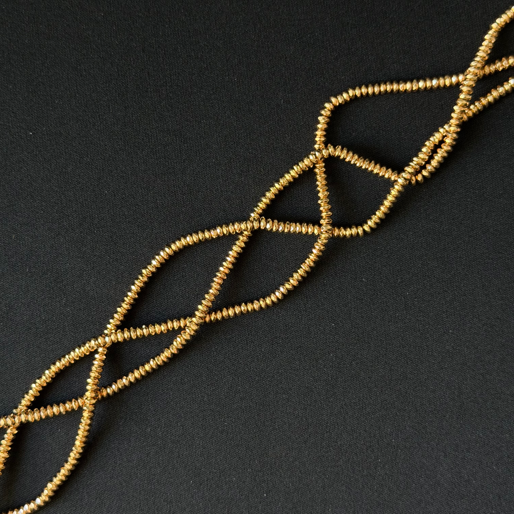 A strand of small gold-colored seed beads arranged in a braided or twisted pattern on a dark, textured background.